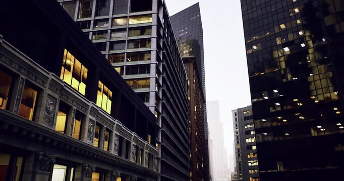 foggy city street between glass skyscrapers, wet pavement reflecting amber office lights, ornate stone facades juxtaposed with sleek steel curtain walls, narrow