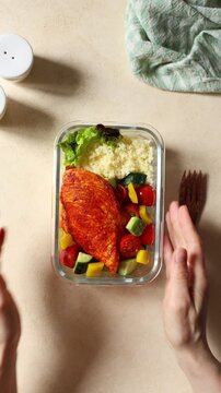 Vertical top view video of hands opening a glass meal prep container with juicy paprika chicken breast, couscous, avocado and fresh vegetables, then eating with a fork.