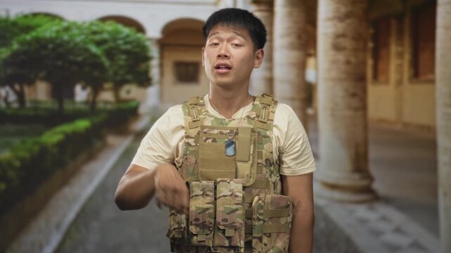 Man in tactical vest and dogtags places hand on chest and points finger in a building corridor lined with columns; duty pride resolve.