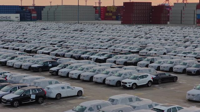Rows of cars at port vehicle terminal with container stacks at golden sunset