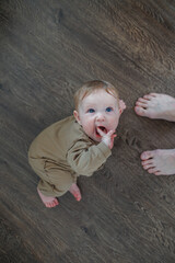 Fototapeta premium High angle shot of a baby sitting on the floor with fingers in mouth near adult feet