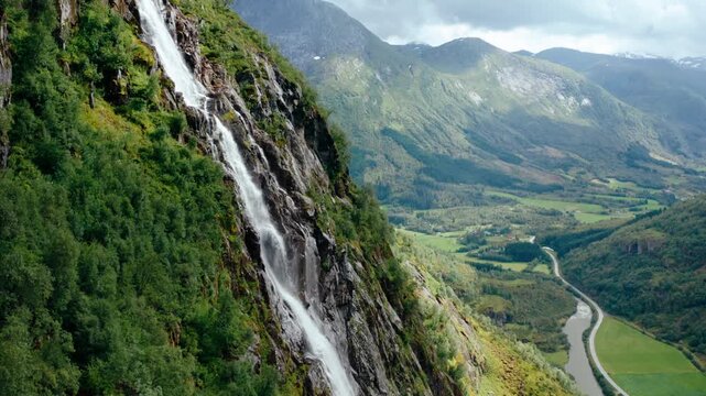 Majestic aerial drone footage of a powerful waterfall cascading into a wild mountain river valley in Klakegg, Norway. Scenic summer wilderness landscape with green peaks captured in summer