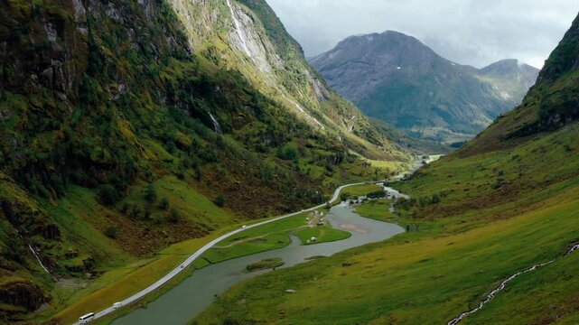 Aerial drone zoom in footage shows vehicles driving on scenic valley road alongside flowing river in Klakegg, Norway. Majestic green mountains landscape captured during sunny July summer day