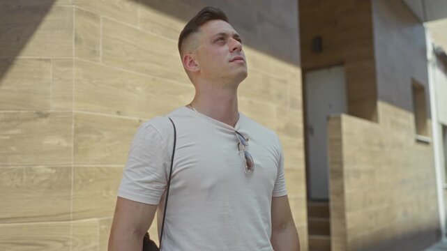 Man with sunglasses hanging from his shirt crosses bare forearms, tilts his head and gazes upward beside a modern wood tiled building entrance while carrying a drawstring bag; calm confidence.