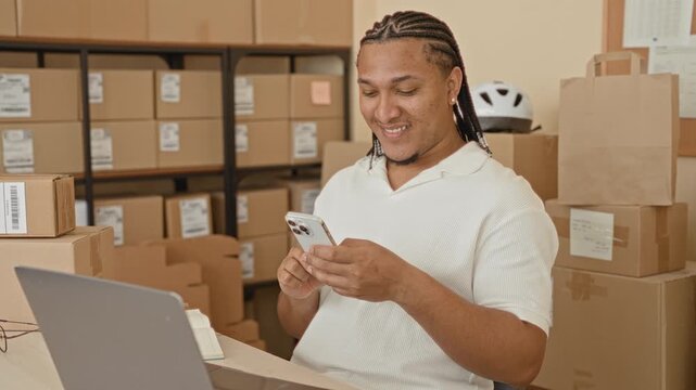 Man smiling and tapping smartphone while seated at laptop amid stacked parcel boxes in warehouse packing area; small business confidence.
