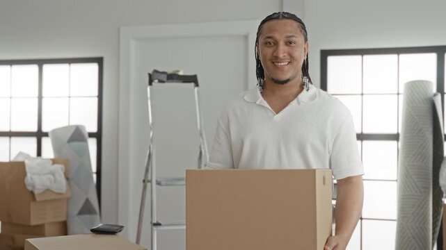 Man smiling and holding a cardboard box at chest in building with ladder and stacked boxes visible; moving day optimism.