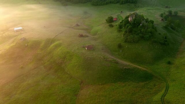 Aerial view of rolling green hills and a rocky outcrop shrouded in morning mist during sunrise in Romania.