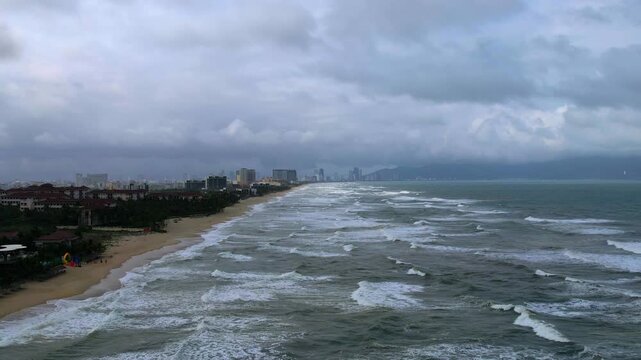 Aerial video of beautiful long sandy beaches of Da Nang in Vietnam overlooking the seaside resorts with huge waves and strong wind on a cloudy moody gray day with city skyline in the background and li