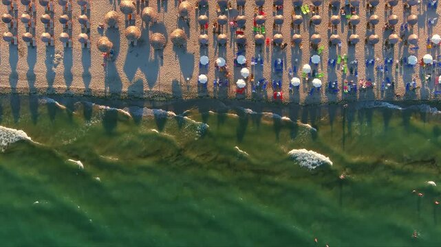 Aerial view of rows of straw umbrellas on a sandy beach with people swimming in the turquoise sea and waves crashing in Paralia, Greece.