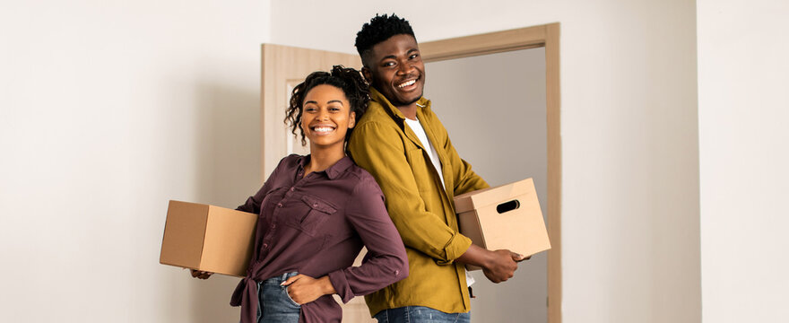 A couple carries brown boxes while smiling in a room. They stand back to back in a newly moved-in space. The sunlight shines through the open door behind them, showing a fresh start.