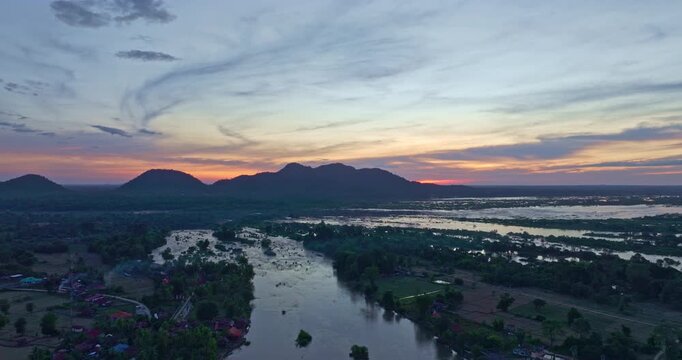 A scenic aerial view of the red bridge crossing the Mekong River near Li Phi Waterfall also known as Tad Somphamit at sunset. The tranquil river landscape with rural farmland and distant mountains