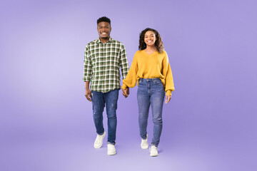 Obraz premium Two people hold hands while walking on a purple background. They appear happy and relaxed. The scene captures a light moment between them. They wear casual clothing and smile at the camera.