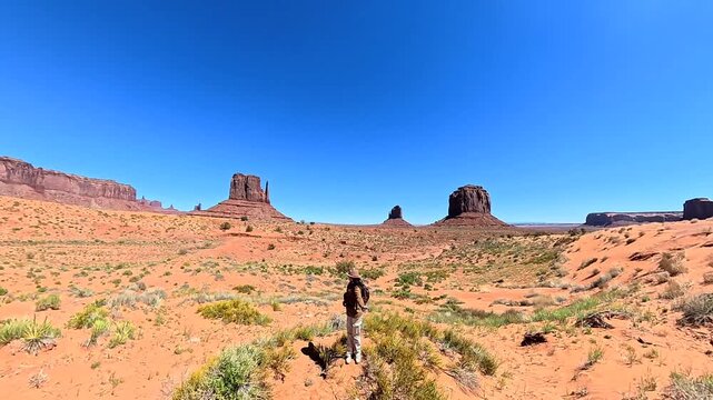 Young female traveler with a cowboy hat and sunglasses joyfully walking backwards, embracing the freedom of her journey through the iconic red sand desert of monument valley, arizona