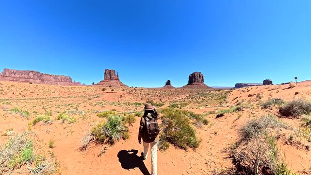 Young female traveler with a backpack and hat walking through the iconic red sand desert of monument valley, admiring the stunning buttes and mesas on a bright, sunny day in arizona