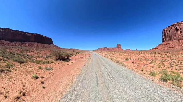 Point of view shot driving on a dirt road through the iconic red sand desert landscape of monument valley, with majestic buttes and mesas under a clear blue sky on a sunny day