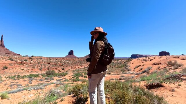 Young smiling woman with a backpack and camera admiring the scenic landscape of monument valley, with the iconic sandstone buttes in the background on a sunny day in the american southwest