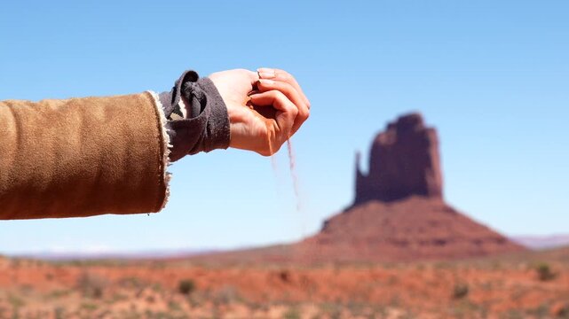 Womans hand sifting red sand through her fingers with the iconic buttes of monument valley in the background, symbolizing the passage of time and a connection with nature