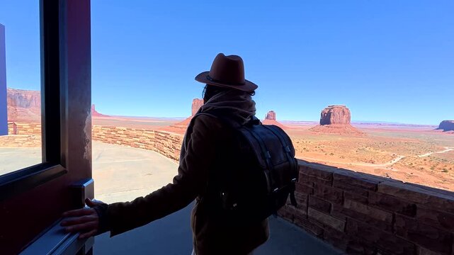 Young female traveler wearing a hat and backpack walking out of a visitor center to admire the stunning panoramic view of monument valley's famous red sandstone buttes in arizona, usa