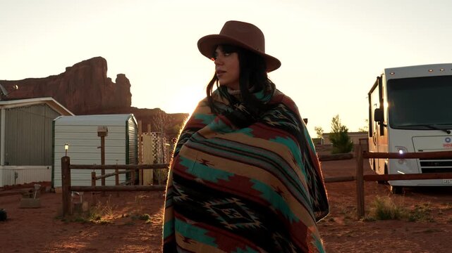 Young adventurous woman wrapped in a traditional patterned blanket standing in the desert of monument valley, arizona, with the warm sunrise glowing behind her and a camper van nearby