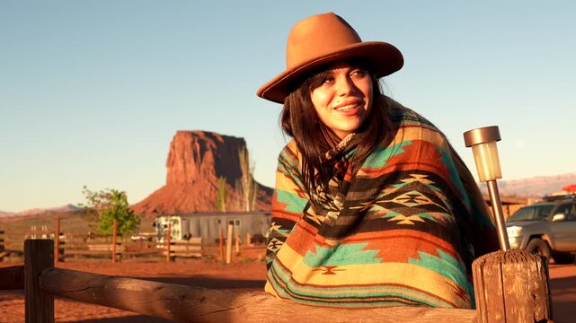 Beautiful young woman wearing a traditional poncho and hat smiling and enjoying the warm sunrise light, with the stunning buttes of monument valley, arizona, creating a picturesque backdrop