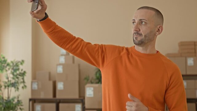 Man in orange sweater taps smartphone with hands amid stacked cardboard boxes at a parcel office building; small business pride.