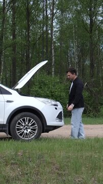 Vertical video man standing by a white stalled vehicle with its hood open, leaning in to inspect the engine, experiencing car trouble on a rural road next to a forest