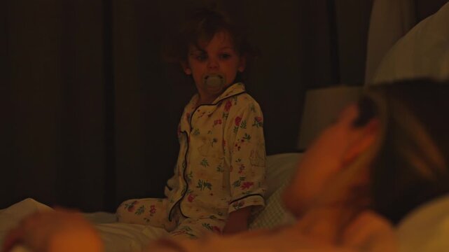Young girl in pajamas with a pacifier sitting in her parents' bed in a dark bedroom at night while her mother sleeps, representing family life, childhood moments, and co sleeping habits