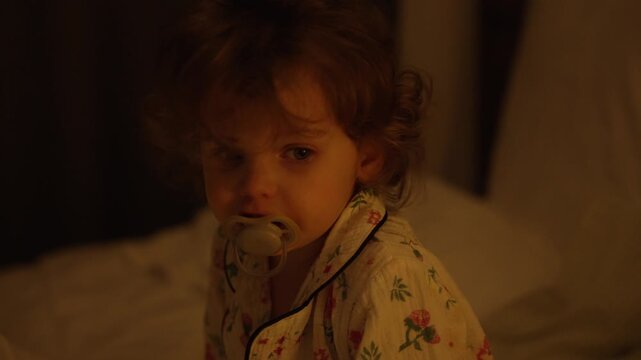 Unhappy toddler girl in floral pajamas crying on her bed in a dimly lit bedroom at night. The upset little child has a pacifier in her mouth and tears in her big sad eyes