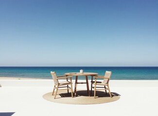 Fototapeta premium Outdoor dining set with two chairs and round table on terrace overlooks blue ocean sea horizon. Minimalist furniture on patio near beach under clear sky.