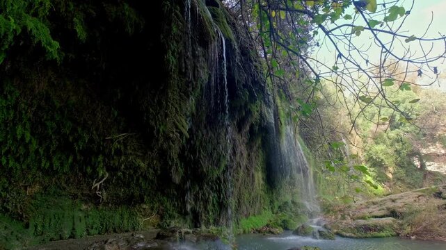 Close-up view of water flowing over mossy rocks in a lush forest, showing a peaceful waterfall scene with green vegetation, wet cliffs and natural daylight for travel and nature footage.