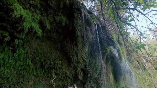 Close-up view of water flowing over mossy rocks in a lush forest, showing a peaceful waterfall scene with green vegetation, wet cliffs and natural daylight for travel and nature footage.