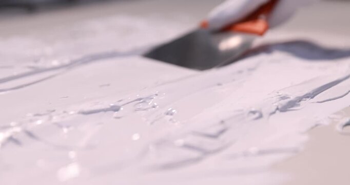 Woman in protective gloves applying white plaster with putty knife onto grey surface, closeup. Camera moving forward