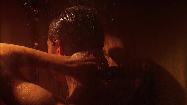 Bare shouldered woman washing her long wet hair under running water in a dark bathroom. The scene, illuminated by warm light, captures a moment of tranquil personal hygiene and daily routine