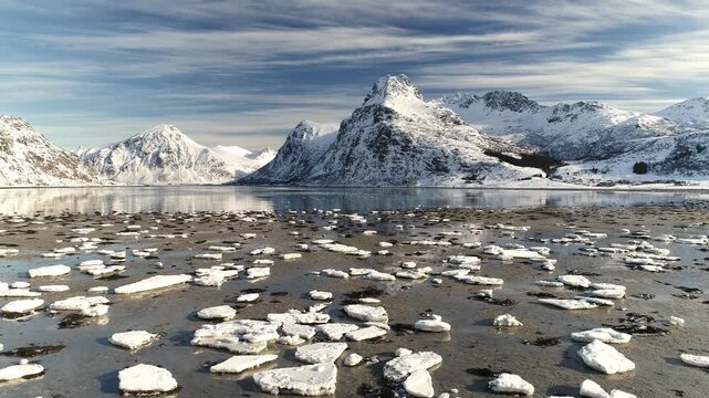 Aerial view of the snow-capped peaks of Lofoten reflecting in the calm water with scattered ice floes under a streaked sky Reine, Nordland, Norway.