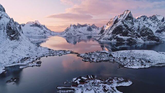 Aerial view of the fishing village of Reine nestled among snow-capped mountains and calm fjords during a pink sunset Reine, Nordland, Norway.