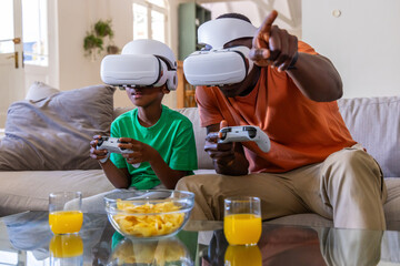 African American father and child playing VR on sofa wearing orange and green holding controllers © wavebreak3