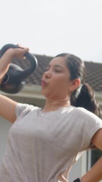 Vertical video: Lifting kettlebell, woman in leggings starting rep to overhead by house for fitness