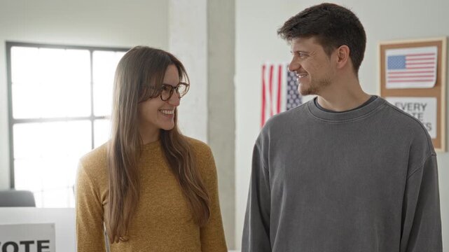 Man and woman smile while making ok hand signs inside a polling building with an american flag and vote booth visible; civic pride.