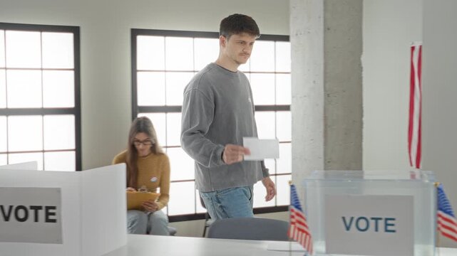 Man places ballot into a clear vote box in a polling building while a woman sits checking a clipboard nearby, us flags and a vote booth visible; civic duty.