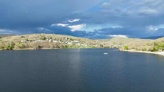 Aerial view of Encoro de Prada reservoir surrounded by green hills and a small village under a dramatic cloudy sky in A Veiga, Ourense, Spain.
