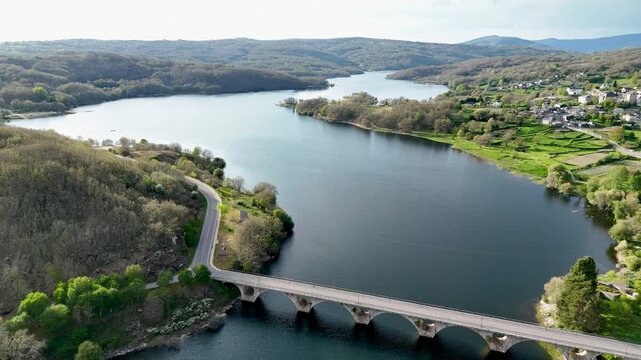 Aerial view of Encoro de Prada reservoir with a stone arch bridge, winding road, and lush green hills under a bright sky A Veiga, Ourense, Spain.