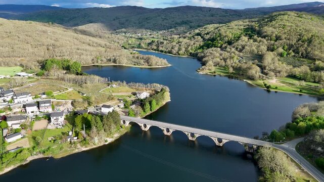 Aerial view of Encoro de Prada bridge spanning the reservoir surrounded by lush green hills and the village of A Veiga, Ourense, Spain.