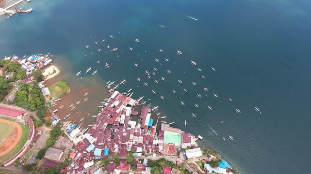 Aerial view of a coastal village with traditional houses and numerous small boats anchored in the blue waters of the bay in Jayapura, Papua, Indonesia.