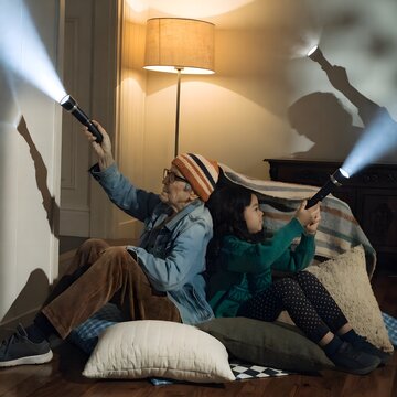 Grandparent and grandchild build a lopsided pillow fort in the living room, flashlight shadows and playful chaos. Cozy family scene for childhood joy.