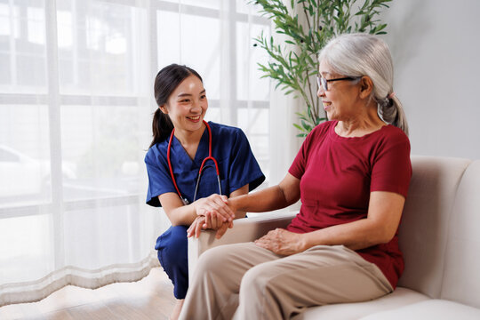 Professional young asian nurse providing caring home care support and medical assistance, holding hands, comforting senior woman patient during a healthcare visit in a domestic environment