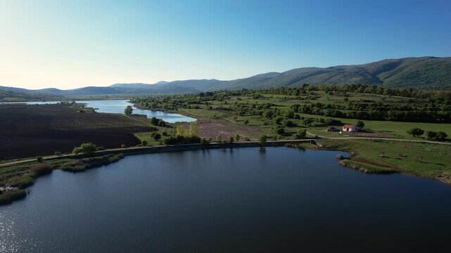 Aerial view of the calm waters of Smilovci Lake with rolling green hills, distant mountains, and a small rural house under a clear blue sky in Smilovci, Pirot District, Serbia.