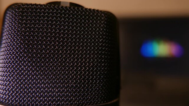 Close-up of a professional podcast microphone. In the blurred background, a laptop displays vibrant, colorful moving audio waveforms. A slow zoom out reveals a minimalist home recording studio setup. 