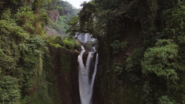 Aerial view of Curug Klenting Kuning waterfall cascading down rocky cliffs surrounded by vibrant tropical vegetation and dense green trees in Kecamatan Limbangan, Central Java, Indonesia.