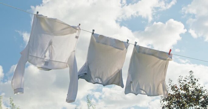 Clean white laundry drying on clothesline in slow motion, breeze blowing fresh clothes under blue sky and white clouds.
