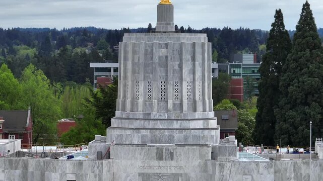 Oregon State Capitol building in Salem featuring golden Oregon Pioneer statue on white marble tower surrounded by lush green trees on overcast day in United States. Aerial rising shot.